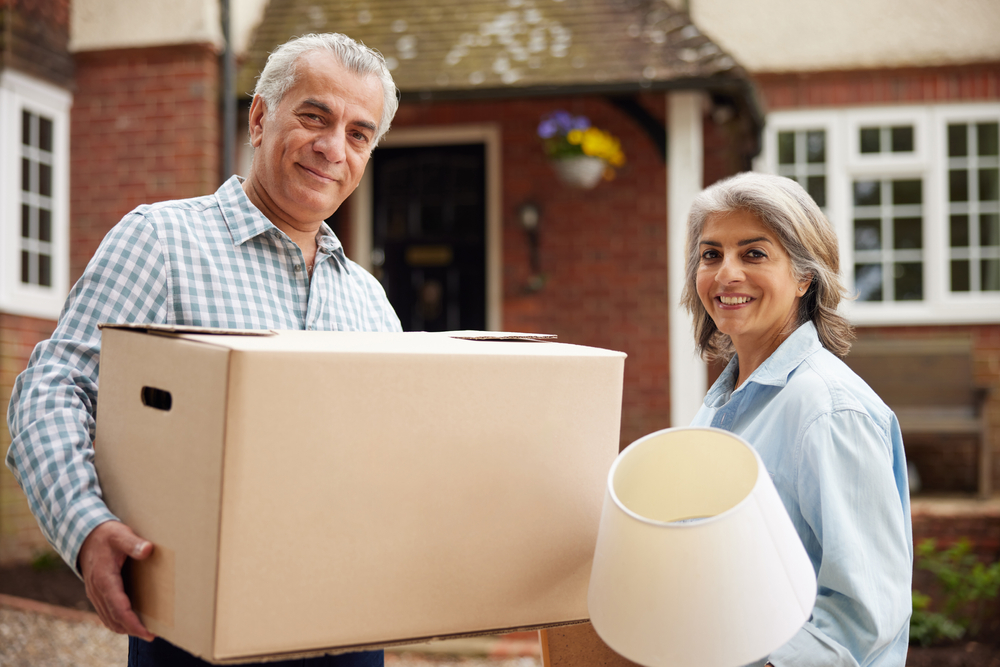 An older couple holding boxes and furniture standing outside a house.