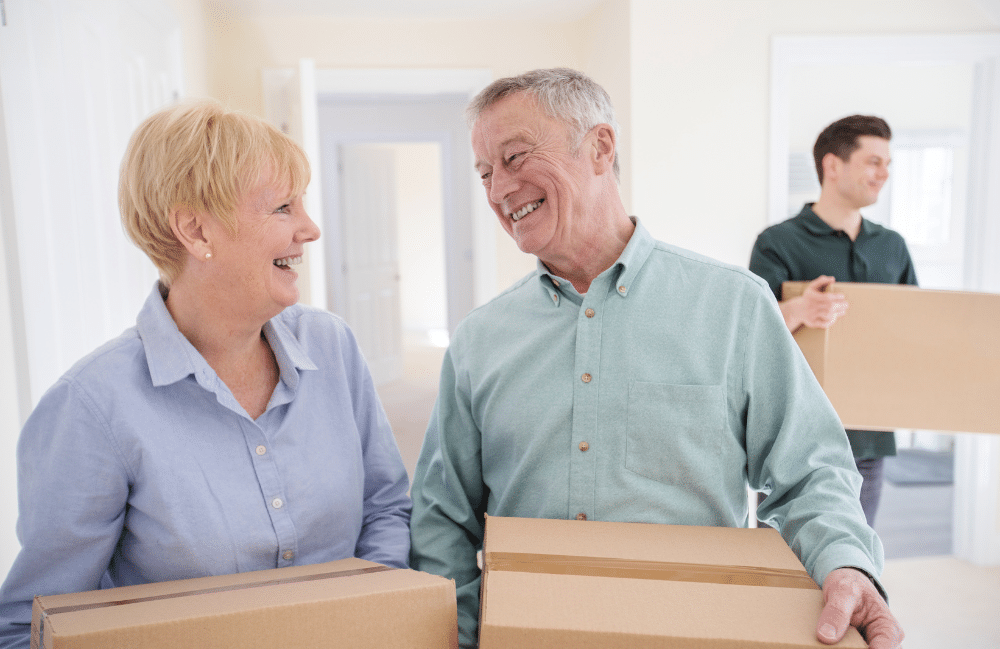 Elderly couple smiling carrying cardboard boxes as a young man walks behind them carrying another cardboard box.