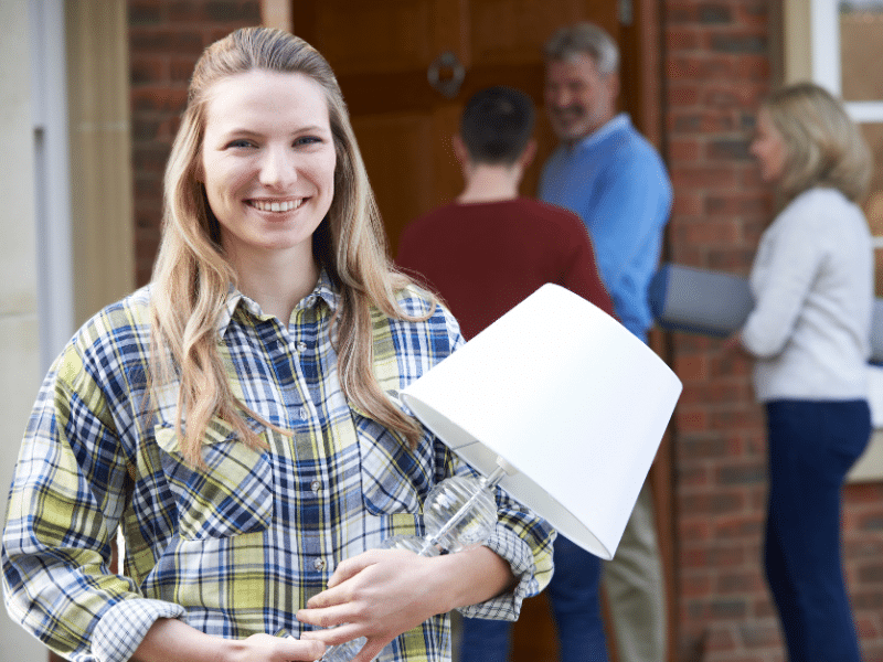Young woman smiling holding a lamp in front of her parents house as her family helps her move out in the background.