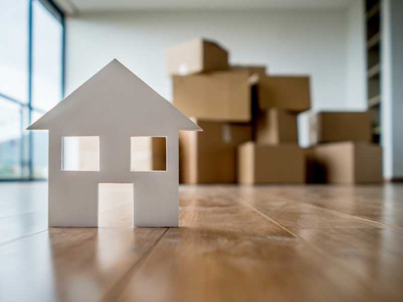 Small cardboard cutout of a house on the floor in front of a pile of cardboard boxes.