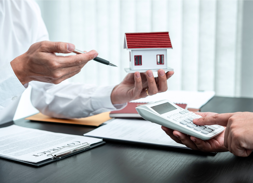 Person holding a calculator talking with someone holding a house miniature and a pen.