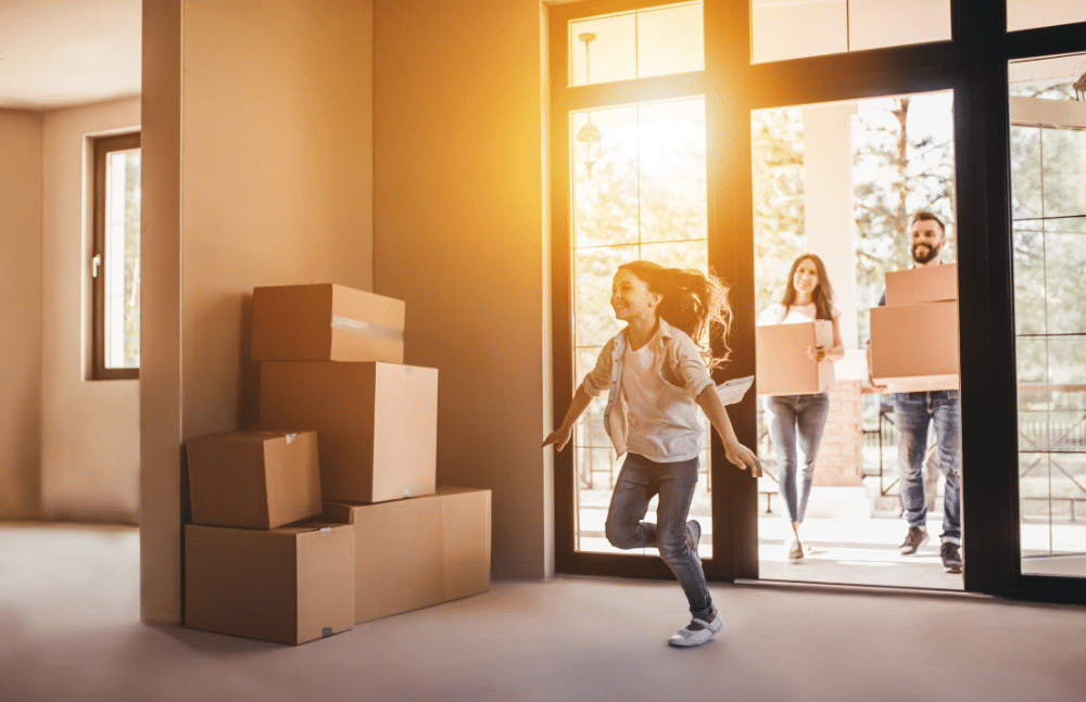 Young girl happily running into her new home as her parents walk in behind her carrying cardboard boxes.